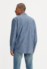 Ljusblå, strukturerad, långärmad chambray-skjorta med knappmanschetter och en böjd nederkant, sedd bakifrån, kombinerad med ljusblå jeans.