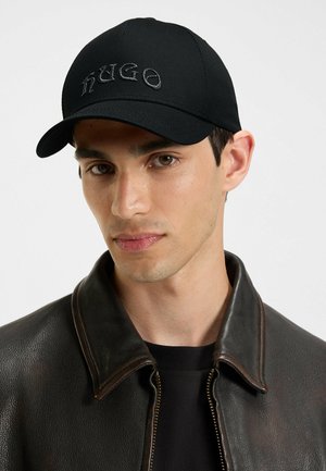 Black cap made of cotton blend with a curved brim; features embroidered "HUGO" in dark thread. Worn with a brown leather jacket.