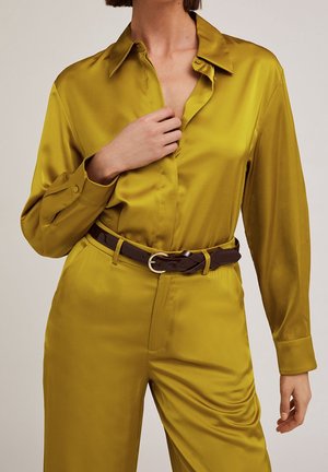 Mujer con camisa de satén amarillo mostaza brillante y pantalones a juego, complementados con cinturón de cuero marrón oscuro con hebilla dorada.