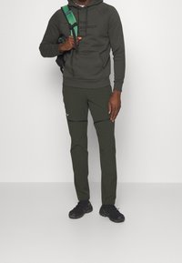 Uomo che indossa un hoodie e pantaloni verde oliva, con uno zaino verde e nero, in piedi contro uno sfondo grigio chiaro uniforme.