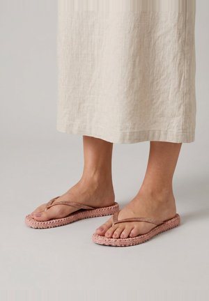 Voeten met roze gebreide teenslippers met dunne bandjes, getoond met een beige linnen rok die onder de knieën eindigt.