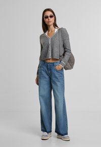 Szürke, kivágott pulóver fehér díszítéssel, világoskék bőszárú farmernadrággal párosítva. A modellen napszemüveg van, és egy szürke steppelt táska van a kezében.