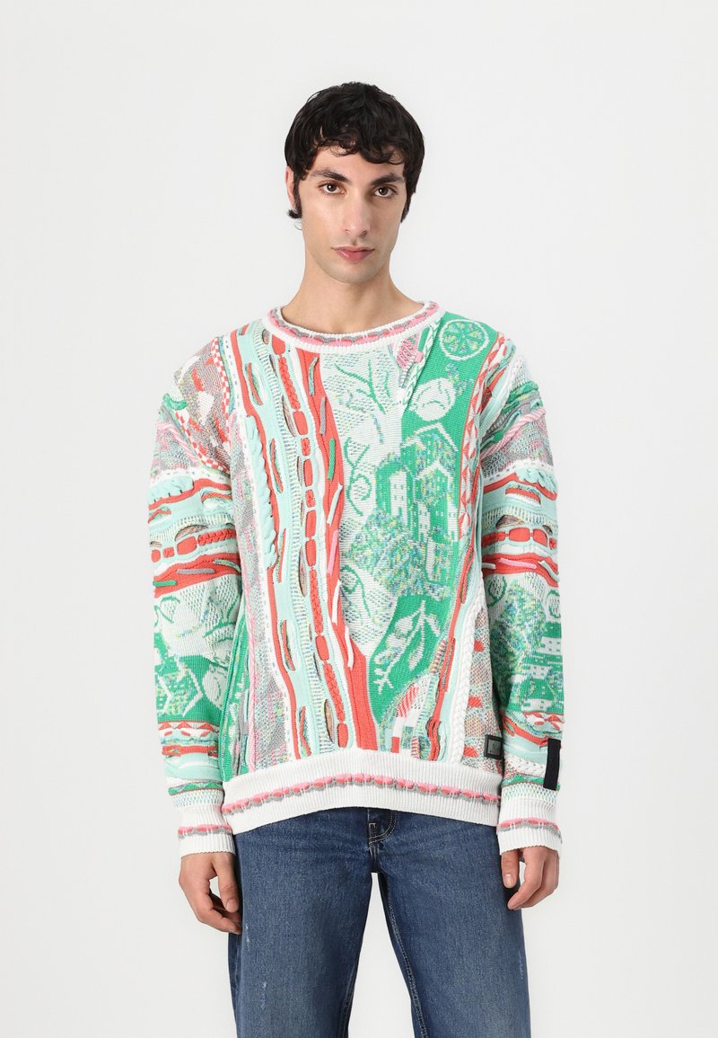 Pull en tricot multicolore avec des motifs verts, rouges et blancs, à encolure ronde et poignets côtelés, porté avec un jean bleu.
