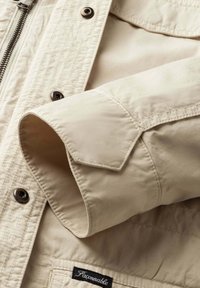 Mouw van een beige jas met gestikte manchetkraag, metalen drukknopen, ritssluiting en een zwart stoffen label met de tekst "Façonnable" op de zak.