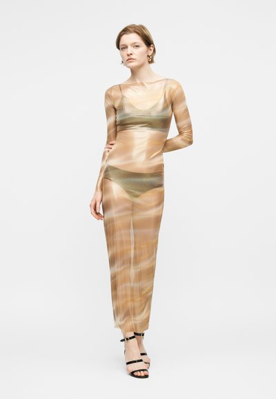 Γυναίκα με κοντά μαλλιά που φοράει ένα διαφανές, μπεζ φόρεμα μακρυμάνικο πάνω από ένα χρυσό μπικίνι, στέκεται με το ένα χέρι πίσω από την πλάτη της.