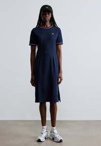 Tumši zils kleita ar īsām piedurknēm, sarkaniem un baltiem apmales elementiem, garums līdz ceļiem ar sānu šķēlumiem, valkāta kopā ar melnu cepuri un baltām sporta kurpēm.