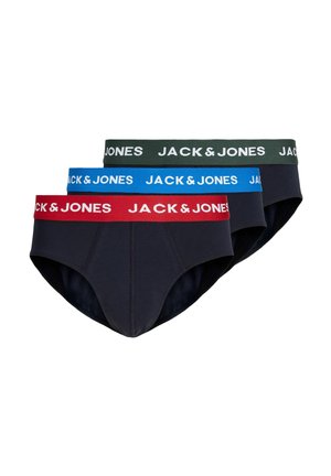 Młody umięśniony mężczyzna z krótkimi blond włosami, ubrany w ciemne majtki z czerwoną gumą z napisem Jack & Jones, stojący na jednolitym tle.