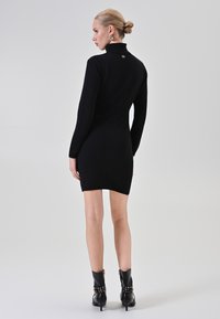 Donna con capelli biondi raccolti in uno chignon basso, che indossa un vestito a maglia colletto alto nero e stivali neri alla caviglia con dettagli metallici, in piedi davanti a uno sfondo uniforme.