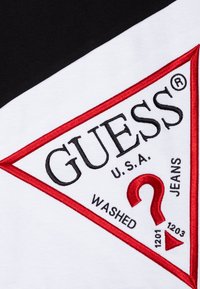 Camiseta blanca y negra con un logo triangular rojo y negro, con el texto "GUESS U.S.A. WASHED JEANS" y un signo de interrogación en el centro.