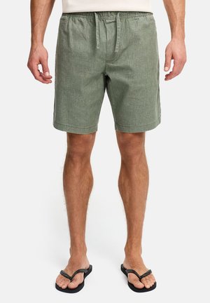 Uomo che indossa pantaloncini verde oliva con coulisse e infradito neri, in piedi su uno sfondo bianco.