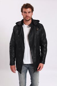 Man med kort brunt hår som bär en svart läderjacka över en vit t-shirt och ljusblå jeans, står mot en neutral bakgrund.