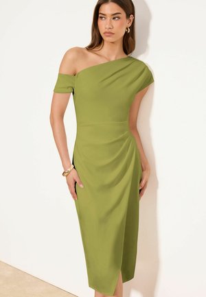 Nő, aki olívazöld egyrészes vállú midi ruhát visel ráncolt derékrészlettel, arany karika fülbevalókkal és arany karkötővel, fehér fal előtt áll.