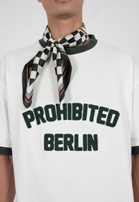 Muškarac u bijeloj majici s tamnozelenim ovratnikom i rukavima, s natpisom "PROHIBITED BERLIN" te šal oko vrata s crno-bijelim karo uzorkom.