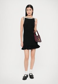 Schwarzes ärmelloses Kleid mit einem Rüschensaum und verziertem Ausschnitt, kombiniert mit schwarzen Schuhen und weißen Socken sowie einer kastanienbraunen Handtasche.