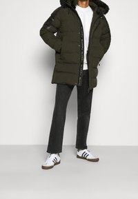 Abrigo acolchado de color verde oliva con capucha desmontable de imitación piel, detalles negros en los hombros, cierre de cremallera y detalles visibles de la marca en la parte delantera.