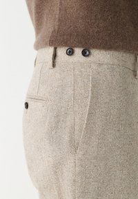 Pantalon beige sur mesure avec une finition texturée, doté de deux boutons noirs à la taille et de poches latérales. Tissu en mélange de coton.