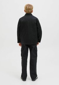 Blond person som står med ryggen mot kameran, iklädd en svart jacka, svarta cargobyxor och svarta sneakers med vita detaljer, på en vit bakgrund.