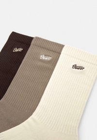 Raidatut sukat tummanruskeana, taupeena ja kermaisen valkoisena, joissa on brodeeratut logo-yksityiskohdat jokaisen parin yläosassa.