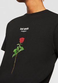 Černé bavlněné tričko s grafickým motivem červené růže se zelenými listy po straně a textem "ztracená mládež" vytištěným nad růží.
