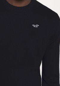 Svart stickad tröja med ribbad halsringning och en liten vit fågel-logotyp på bröstet. Tyget har en slät yta.