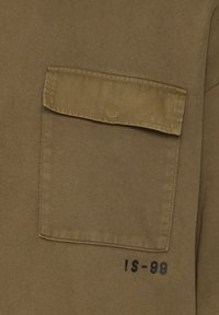 Primer plano de una camisa de tela caqui con un bolsillo de solapa rectangular y texto negro que dice "IS-99" debajo del bolsillo en el lado derecho.