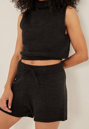 Ärmelloses, schwarzes, gestricktes Croptop und passende schwarze, gestrickte Shorts mit Kordelzug, getragen von einer Person mit lockigem Haar.