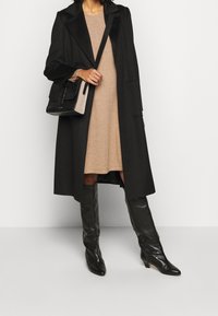 Manteau noir long avec un col cranté, porté sur une robe beige. Associé à des bottes noires montantes et un sac à main texturé noir et beige.