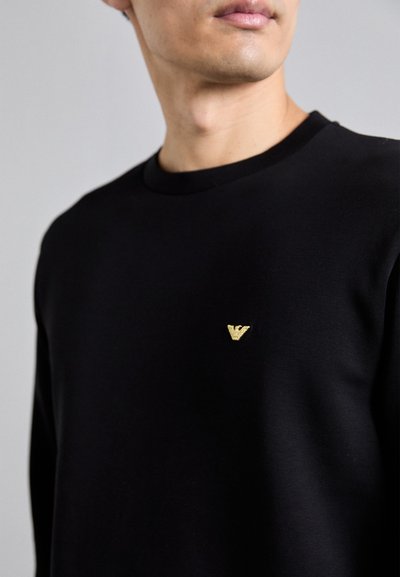 Black sweatshirt with a round neckline and long sleeves, featuring a small gold embroidered logo on the chest and a smooth fabric texture.