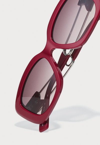 Óculos de sol oversized vermelhos com uma armação de plástico brilhante, lentes escuras em degradê e detalhes em metal prateado nas dobradiças. Forma quadrada com bordas arredondadas.
