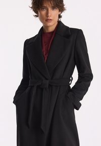 Kobieta w czarnym płaszczu z paskiem, nosząca burgundowy top, stoi z rękami w kieszeniach płaszcza na jednolitym jasnym tle.