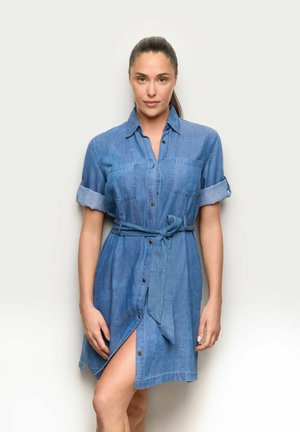 Frau steht vor einem weißen Hintergrund und trägt ein blaues Jeans-Hemdkleid mit hochgekrempelten Ärmeln und einem gebundenen Taillengürtel.