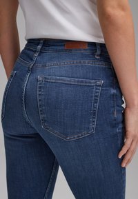 Gros plan d'une personne portant un jean en denim bleu avec des poches arrière visibles et une étiquette de marque en cuir marron, associé à un haut blanc.