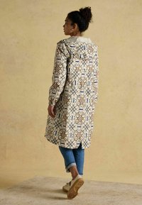 Krēmisks garš mētelis ar ziliem un brūniem rakstiem, ar kapuci, elastīgām piedurknēm un atvērtu apakšu, apvienots ar džinsiem un sporta apaviem.