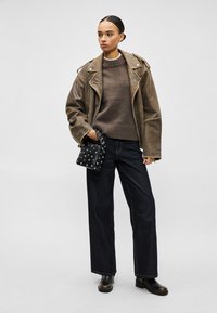 Καφέ δερμάτινο μπουφάν, σκούνο πλεκτό πουλόβερ, τζιν παντελόνι με φαρδιά μπατζάκια σε σκούρο χρώμα, μαύρη τσάντα με ασημένιους τρούκ, μαύρα μποτάκια αστραγάλου, ουδέτερο φόντο.