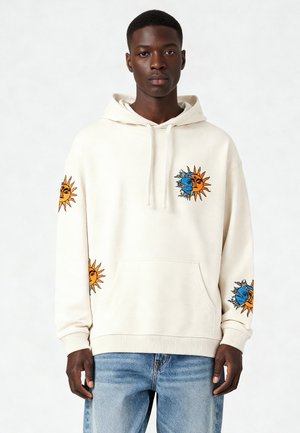 Sweat à capuche crème avec des graphiques de soleil et de lune sur les manches, doté d'une poche kangourou et d'un cordon de serrage. La texture semble douce et décontractée.