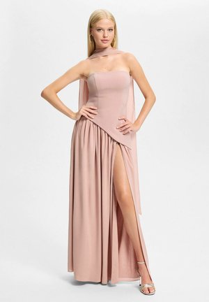 Blondynka stojąca z rękami na biodrach, ubrana w różową suknię na ramiączkach z wysokim rozcięciem oraz pasującą przezroczystą szarfę.