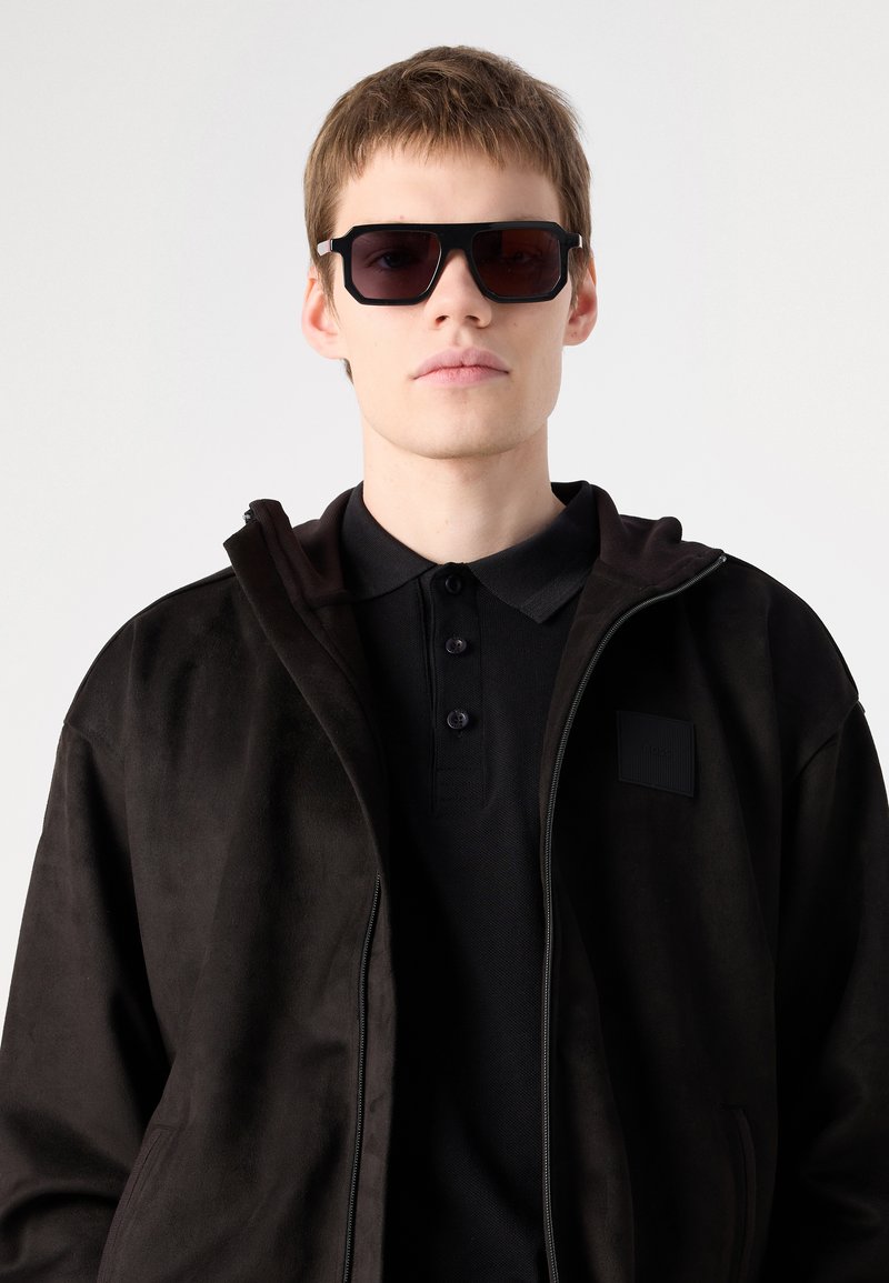 Schwarze Wildlederjacke mit Reißverschluss vorne, kombiniert mit einem schwarzen Polohemd. Dunkle, rechteckige Sonnenbrille. Glatte Textur, minimalistisches Design.