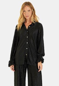 Camisa negra de manga larga hecha de una tela texturizada con un patrón de diamantes, que cuenta con un cierre de botones y puños fruncidos.