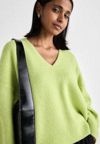 V-veida, pārspīlēts džemperis lime zaļā krāsā, izgatavots no mīkstas adītas auduma. Iezīmē garas piedurknes un ribbētu apakšmali, kopā ar spīdīgu melnu soma siksnu.