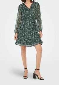 Donna che indossa un abito floreale con maniche lunghe e scollatura a V, lungo fino al ginocchio, abbinato a sandali con tacco blocco e cinturino alla caviglia neri.
