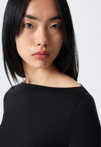 Záber zblízka osoby nosiacej čierne priliehavé tričko s lodičkovým výstrihom. Materiál vyzerá hladký, s rovnými lesklými čiernymi vlasmi.