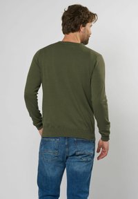 Jersey verde de manga larga con un cuello y dobladillo acanalados, combinado con jeans azules. La tela tiene una apariencia texturada y una forma ajustada.