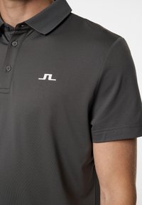 Mörkgrå pikétröja i slätt tyg, med spetsig krage, tre knappar och en vit logotyp på vänster bröst.