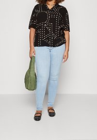 Svart kortärmad topp med beige geometriskt mönster, ljusblå smala jeans, grön kviltad handväska och svarta balettklackar med nitar.