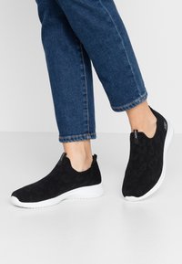 Mustahdetut mustat liukupäiväsaappaat, joissa on teksturoitu kuvio, valkoiset pohjat ja matala leikkaus, yhdistettynä tummiin farkkuihin.