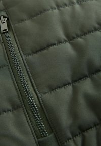 Grüne Pufferjacke aus Stoff mit gestepptem Muster und sichtbarem Reißverschlussdetail. Verfügt über eine glatte Oberfläche und einen kontrastierenden Reißverschluss.