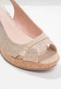 Sandales compensées beige avec un dessus texturé scintillant et une plateforme en liège. Le design à bout ouvert présente des sangles croisées et une doublure intérieure douce.