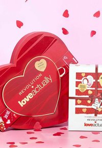 Röd hjärtformad adventskalender med guldakcenter, med texten "Revolution loveactually." Intill en vit presentförpackning med röda ränder och band.