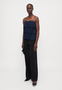 Navy pleated top with three layers, thin straps, paired with black wide-leg trousers and black pointed shoes with metallic accents.