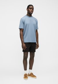 Homme debout portant un t-shirt à manches courtes bleu clair, un short noir, des chaussettes blanches et des baskets marron sur un fond blanc uni.